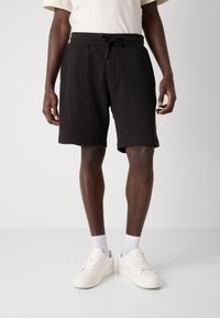 Shorts neri con texture a waffle, dotati di cintura elastica e lacci, con tasche laterali e vestibilità comoda, abbinati a sneakers bianche.