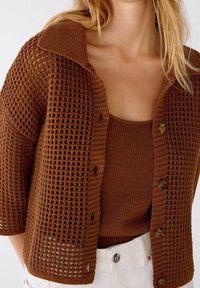 Hnědý pletený kardigan s volným proplétáním, krátkými rukávy a velkým límcem; má dřevěné knoflíky a je vrstvený přes sladěný top.