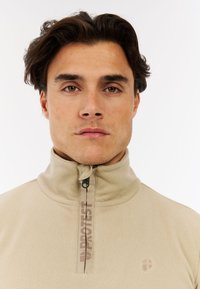 Beige fleece pullover med stående krave, halv lynlås og markant logo-detalje på brystet. Blød tekstur, minimalistisk design og enkle accenter.