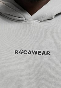 Lysegrå hættetrøje med et sort "RCAWEAR" logo på brystet, lavet af blødt stof med en glat tekstur og med snoretræk i hætten.