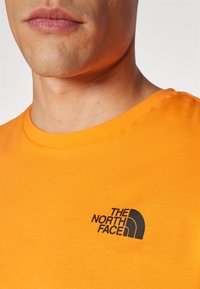 Oranžinė medvilninė marškinė su juodu "The North Face" logotipu kairėje krūtinės pusėje. Apvali iškirptė, trumpomis rankovėmis, lygios tekstūros.