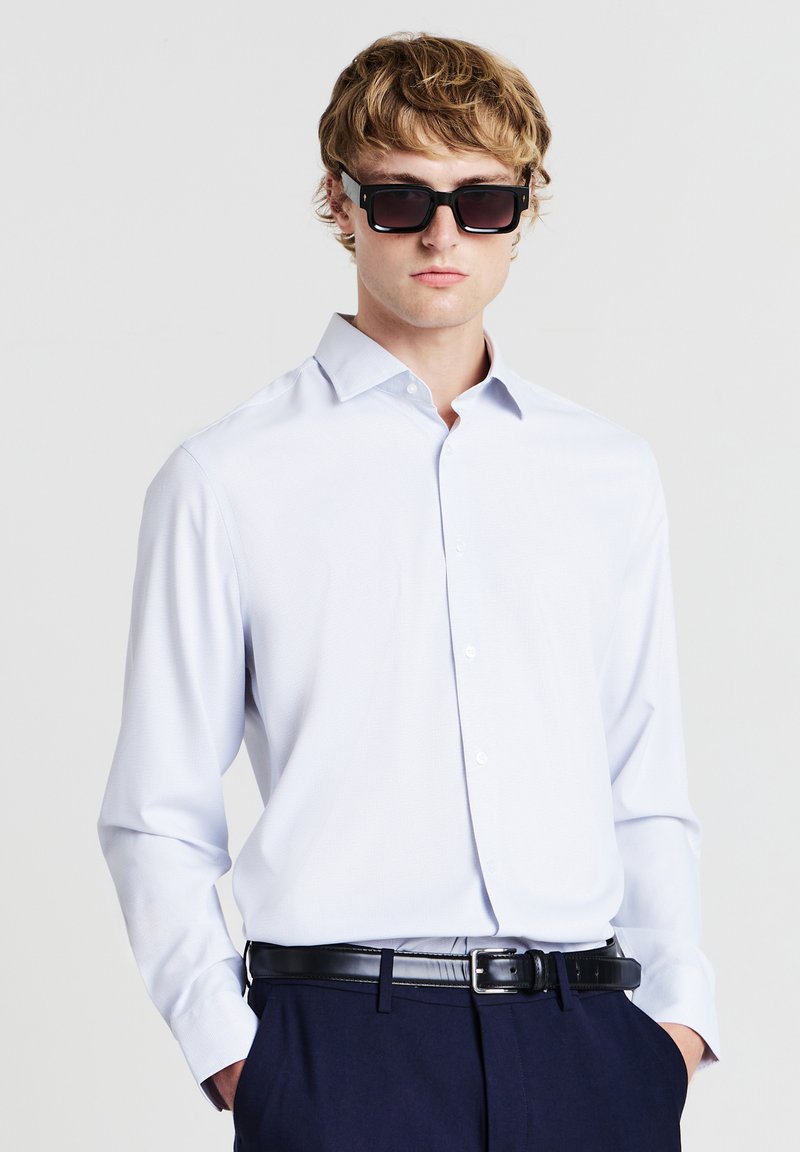 Giovane uomo che indossa occhiali da sole neri oversize, camicia azzurro chiaro con bottoni, pantaloni scuri e cintura in pelle nera, posando con le mani in tasca.