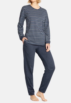 Vrouw die een marineblauw gestreept shirt met lange mouwen en effen marineblauwe broek draagt, staat op blote voeten met één hand in de zak en de andere rustend op de dij.