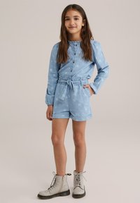 Világoskék farmer rövid overall virágmintával, redőzött hosszú ujjakkal, gombos záródással és illeszkedő anyagövből. Bézs katonai csizmával viselve.