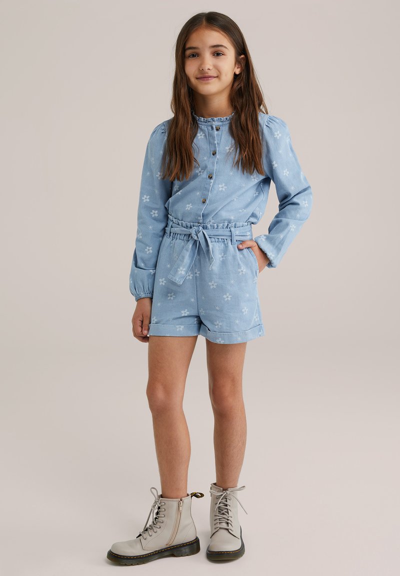 Világoskék farmer rövid overall virágmintával, redőzött hosszú ujjakkal, gombos záródással és illeszkedő anyagövből. Bézs katonai csizmával viselve.