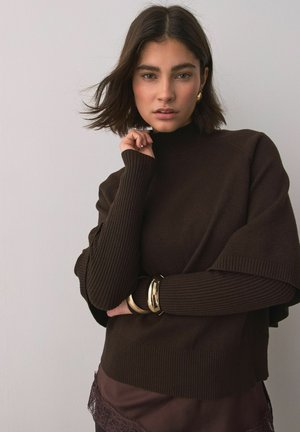 Woman with short brown hair wearing a dark brown turtleneck sweater, gold bracelets, and earrings, posing with hand near face against light wall.