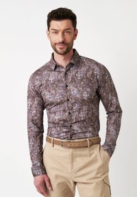 Bruine shirt met een paisley patroon, knoopsluiting aan de voorkant en lange mouwen. Gecombineerd met beige broek en een brede bruine riem.