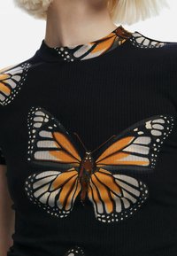 Camiseta negra de canalé con grandes impresiones de mariposas en naranja, amarillo y blanco. El diseño incluye patrones de alas detallados y acentos.