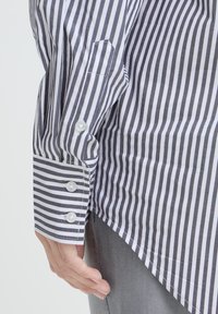 Striped shirt features narrow black and white vertical stripes, a buttoned cuff, and a curved hem, made from lightweight cotton fabric.