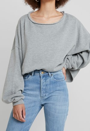 Nainen pukeutuneena ylisuuruneen vaaleanharmaan hupparin höyhenhihalla ja vaaleansinisiin, korkeavyötäröisiin farkkuihin, seisomassa yhdellä kädellä lanteilla.