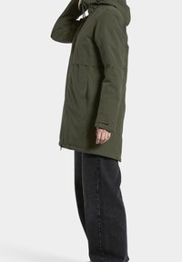 Parka verde oliva con cremallera frontal, capucha ajustable y puños elásticos, llevada sobre pantalones negros de pierna ancha. Tejido suave con acabado mate.