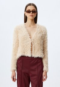 Faux-Fur-Jacke in hellem Beige mit tiefem V-Ausschnitt, langen Ärmeln und verkürztem Design. Kombiniert mit weinroten Hosen.
