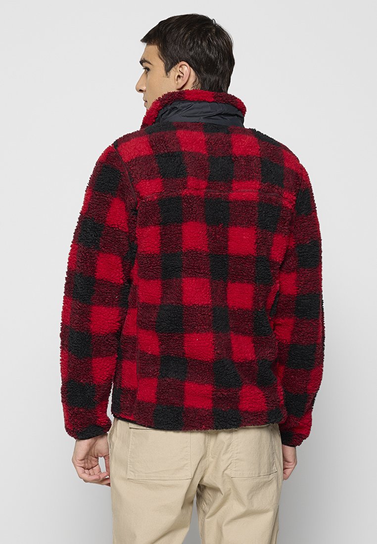 Czerwono-czarna, w kratę polarowa kurtka z wysokim kołnierzem, miękka w dotyku, z zamykaną na zamek kieszenią na plecach. Model odwrócony plecami.
