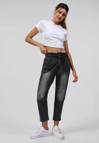 Kvinne iført en hvit kort t-skjorte, høyt liv svarte pants med snor, og hvite joggesko, poserer foran en ensfarget bakgrunn.