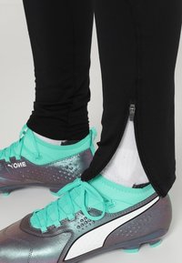 Svarta träningbyxor med ankeldragkedjor och en åtsittande design, kombinerat med turkosa och grå fotbollsskor med en texturerad yta.