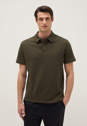 Homme portant un polo à manches courtes vert olive et un pantalon noir, debout avec une main dans la poche sur un fond uni.