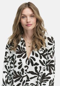 Blus putih dengan pola daun hitam besar, desain tanpa kerah, lengan panjang, dan plakat berbutang. Kalung liontin emas menonjolkan.
