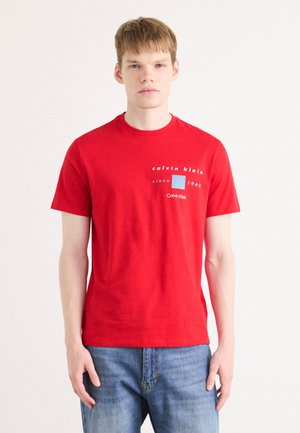 Camiseta de algodón roja con mangas cortas, que presenta texto blanco y un gráfico cuadrado azul. Combinada con unos vaqueros de mezclilla azules.