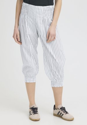 Kvinde iført hvide korte bukser med lodrette sorte striber og elastiske manchetter, kombineret med beige sneakers med sorte striber.