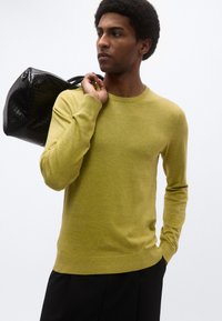 Πλεκτό πουλόβερ σε ελιά χρώμα, στρογγυλή λαιμόκοψη, μακριά μανίκια, λείο ύφασμα. Το μοντέλο κρατά μια μαύρη πλεκτή τσάντα ώμου.