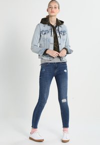 Light blue denim jacket with pockets, gray sweatshirt, camouflage hood, dark blue skinny jeans with ripped details, white sneakers with pink accents.