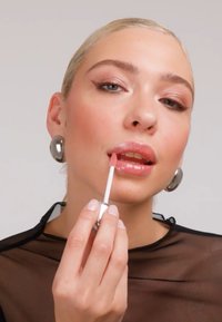 Femme aux cheveux blonds plaqués en arrière portant de grandes boucles d'oreilles argentées applique un gloss à lèvres rose, vêtue d'un haut noir transparent sur un fond uni.