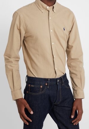 Uomo che indossa una camicia beige con bottoni e maniche lunghe e jeans blu scuro, in piedi con le mani vicino alle tasche su uno sfondo semplice.