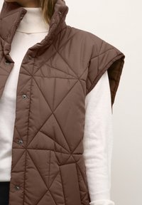 Brązowa, pikowana kamizelka bez rękawów z wysokim kołnierzem i zapięciem na napy, noszona na białej bluzce z długim rękawem. Tekstura jest gładka i lekko wypełniona.
