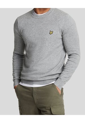 Mężczyzna ubrany w jasnoszary sweter typu crewneck z żółtym logo ptaka, pod którym ma białą koszulę, oraz oliwkowe zielone spodnie typu cargo, z rękami w kieszeniach.