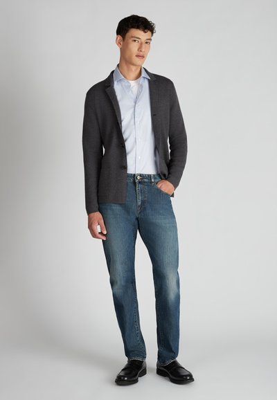 Grijze cardigan over een lichtblauw shirt, gecombineerd met vervaagde blauwe jeans en zwarte schoenen. De cardigan heeft een knoopsluiting en zakken.