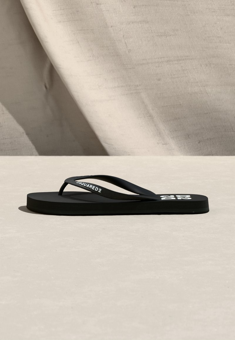 Sandale tong noire à semelle plate, avec une lanière de marque et un logo blanc imprimé sur la semelle intérieure, sur un fond en tissu beige.