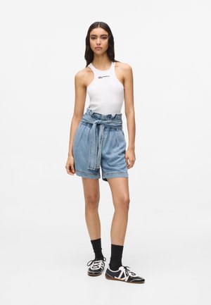 Kobieta stojąca, ubrana w białą, bezrękawnikową bluzkę, jeansowe szorty z wysokim stanem i paskiem w stylu paperbag, czarne skarpetki oraz czarno-białe tenisówki.