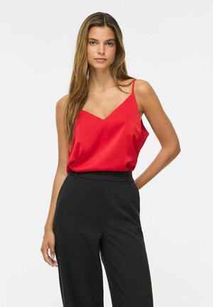Femme debout devant un fond uni, portant un haut rouge sans manches à fines bretelles et un pantalon noir taille haute, regardant devant elle.