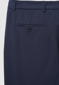 Pantalon bleu marine en tissu lisse, avec une poche arrière unique dotée d'un bouton noir, coupe ajustée et plis marqués.