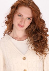 Cardigan crema a intrecci cavo con scollatura a V, caratterizzato da tre grandi bottoni dorati, indossato sopra un top a coste beige chiaro. Morbido al tatto e caldo.