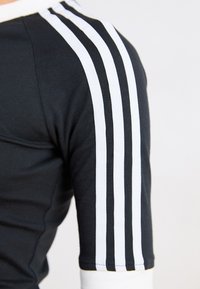Svart atletisk topp med tre hvite striper nedover ermene, rund hals, og en tettsittende design med strukturert stoff.