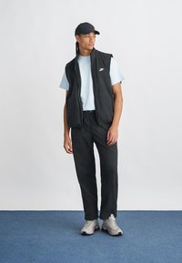 Μαύρο αμάνικο γιλέκο πάνω από ανοιχτό μπλε μπλουζάκι, συνδυασμένο με μαύρο παντελόνι και γκρι αθλητικά παπούτσια. Περιλαμβάνει το λογότυπο της Nike στο γιλέκο.