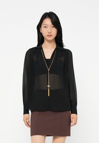 Nainen, jolla on päällään läpinäkyvä musta pitkähiha blouse, kultaketju, jossa on riippuvat hapsut, ja ruskea hame, taustanaan tasainen valkoinen tausta.