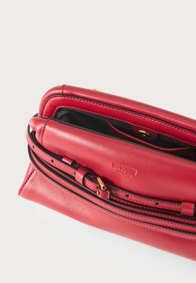 Κόκκινη δερμάτινη τσάντα Moschino χιαστί με ρυθμιζόμενο λουρί, ανοιχτό πάνω μέρος που δείχνει μαύρο εσωτερικό και χρυσό κλείσιμο με κουμπί.