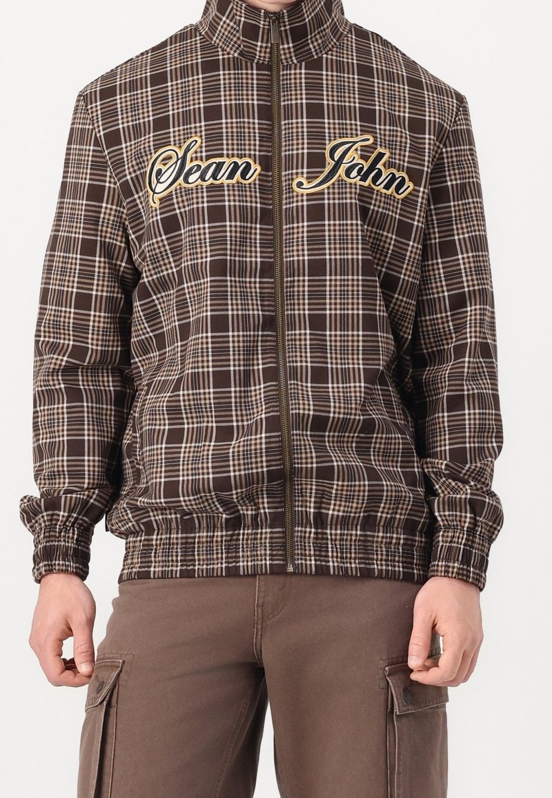 Veste zippée à carreaux marron avec des poignets et un ourlet élastiques, arborant "Sean" et "John" brodés en jaune sur la zone de la poitrine.