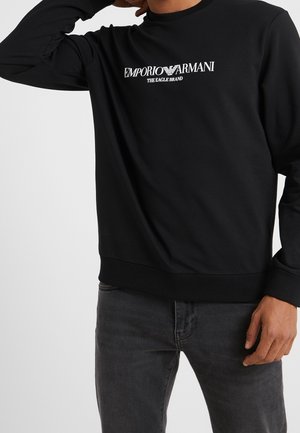 Mężczyzna noszący czarną bluzę Emporio Armani z białym logo na piersi oraz ciemnoszare dżinsy na jednolitym tle.