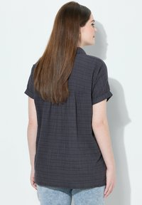 Camisa de manga corta de color gris oscuro con una tela texturada, que presenta un pliegue en la parte posterior y mangas arremangadas, combinada con jeans de color azul claro.