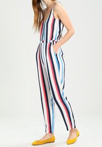 Randig jumpsuit i vitt, marinblått, rött och blått. Har en v-ringning, är ärmlös och har ett elastiskt midjeband. Fickor på sidorna.