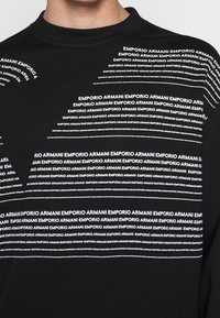 Svart sweatshirt med upprepande vit text "Emporio Armani" i olika längder och orienteringar, med rund halsringning och lång ärm.