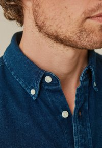 Vue partielle du cou et de la mâchoire d’un homme avec une légère barbe, portant un col de chemise en denim bleu foncé boutonné.