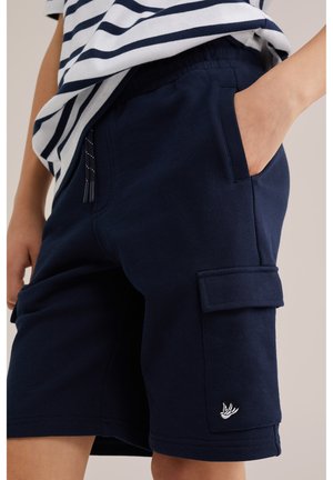 Barn iført marineblå cargoshorts med en lille hvid fuglelogo på lommen og en stribet skjorte, der er stukket ind.