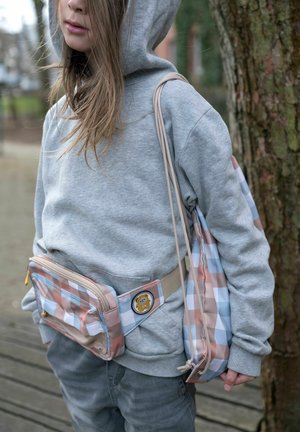 Ung person i grå hættetrøje, iført matchende ternet bæltetaske og rygsæk med snøre samt tegneseriepatch, stående nær et træ udendørs.