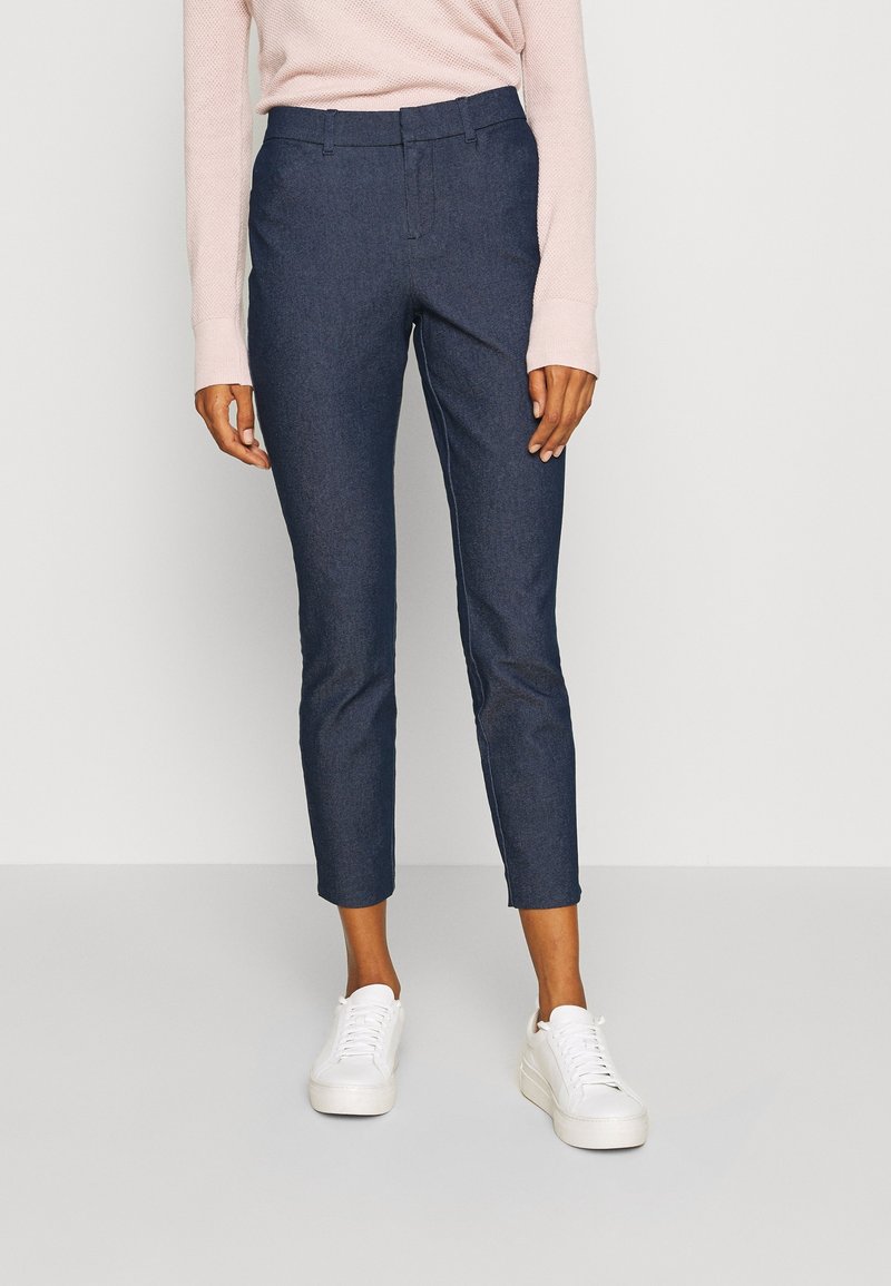 Personne portant un pantalon bleu marine slim fit longueur chevilles, une chemise rose pâle à manches longues et des baskets blanches, debout devant un fond uni.