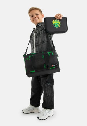 Chłopiec w czarnym dżinsowym stroju trzyma czarną i zieloną torbę LEGO Ninjago oraz pasujący do niej śniadaniówkę z napisem „Ninja Training”.