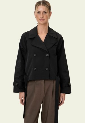 Frau trägt eine schwarze, zweireihige, kurz geschnittene Jacke mit weiten Revers und braune, hoch taillierte Hose, steht vor einem neutralen Hintergrund.